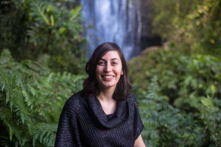 A woman with shoulder-length dark hair and a black sweater stands in front of green foliage, smiling at the camera. A waterfall is visible in the blurred background.