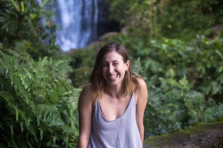 A woman with shoulder-length brown hair and a gray tank top stands outdoors, smiling, with lush green plants and a waterfall in the background.