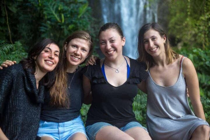 Four women are sitting close together outdoors, smiling at the camera. They are surrounded by lush green foliage, with a waterfall visible in the background.