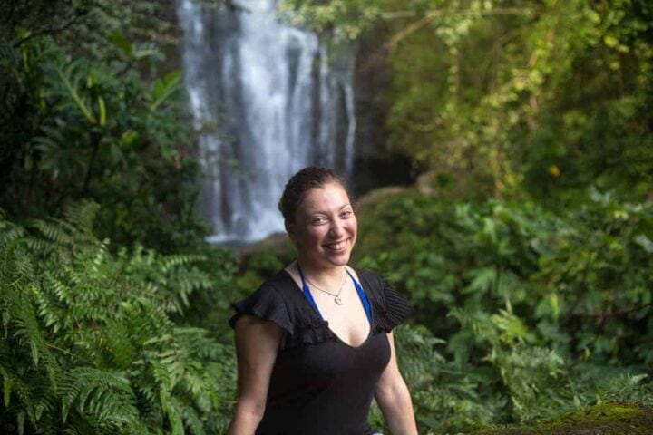 A woman stands smiling in front of a waterfall surrounded by lush greenery. She is wearing a black shirt and a necklace, and her hair is pulled back. The setting appears to be a tropical or forested area.