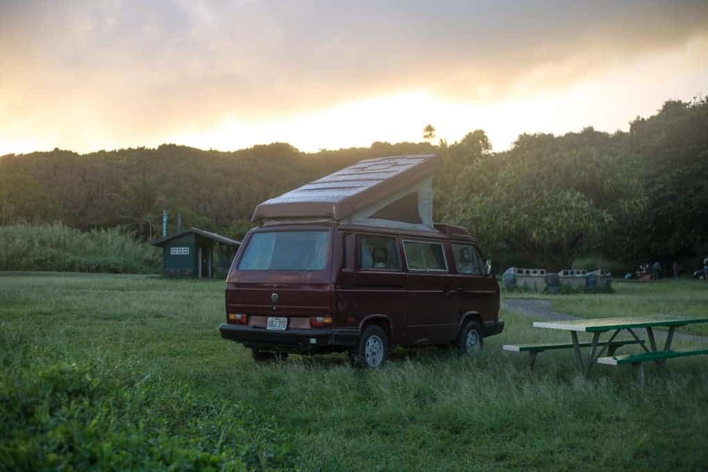 A maroon camper van with a raised roof is parked on grass near a picnic table at a campsite. Trees, a building, and trash bins are in the background under a cloudy sky at sunset.