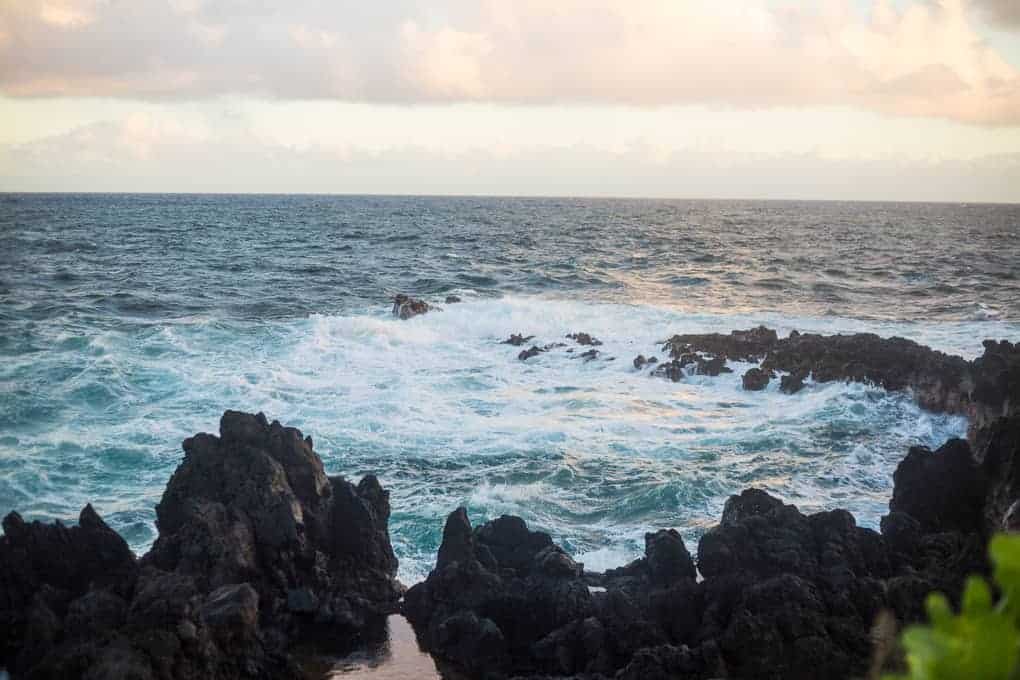 Waves crash against dark, jagged rocks on a coastline under a cloudy sky, with the sun casting a soft light over the ocean and horizon.