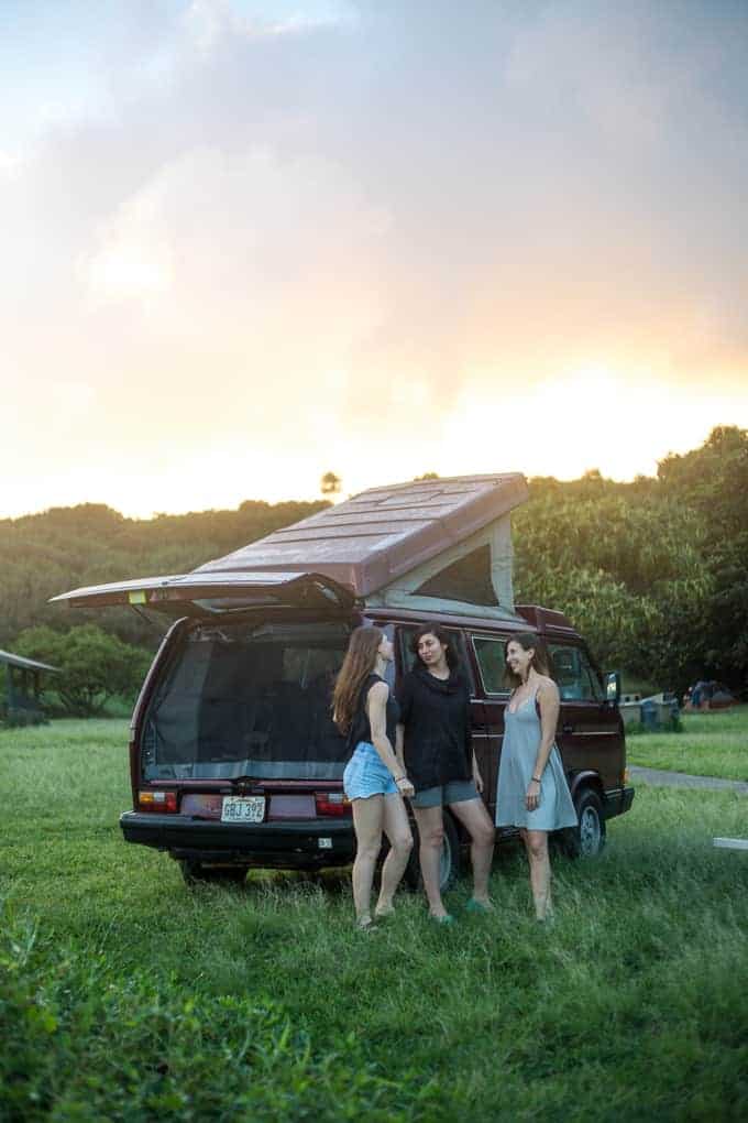 Three people stand and talk next to a maroon camper van with its back door and pop-up roof open, parked on grass in a field at sunset with trees and hills in the background.