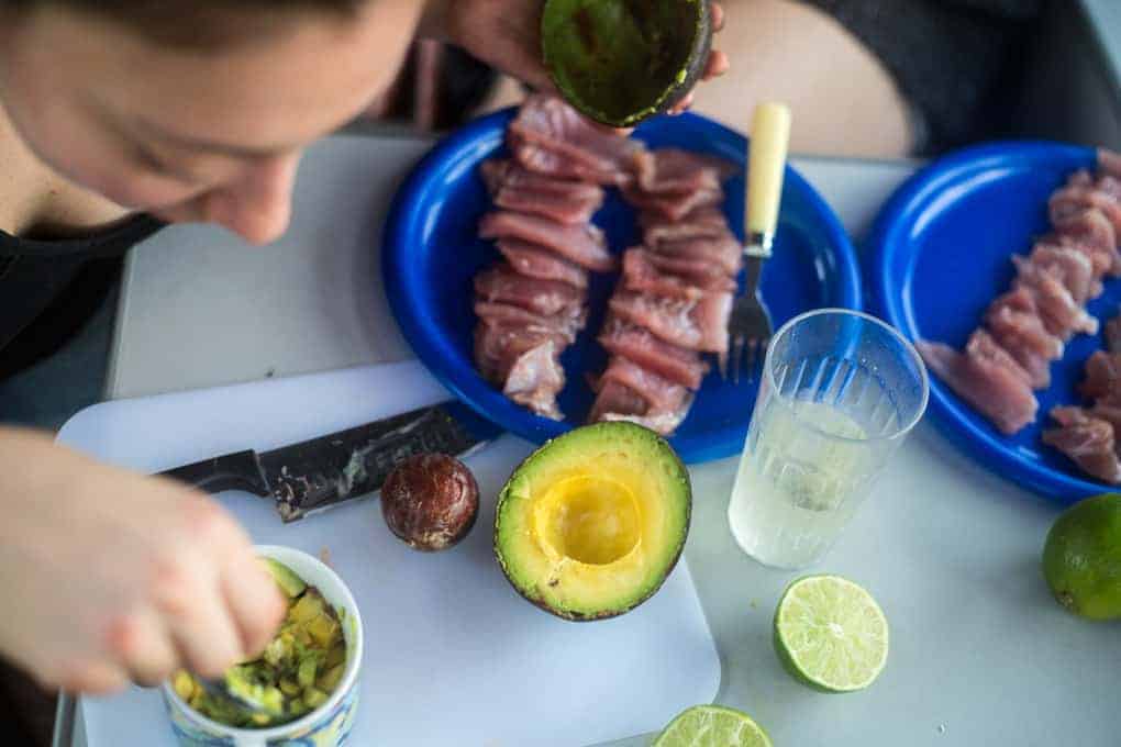 A person prepares food at a table with raw fish pieces on blue plates, a halved avocado, limes, a knife, a plastic cup of water, and a bowl with a spoon. The scene is viewed from above.