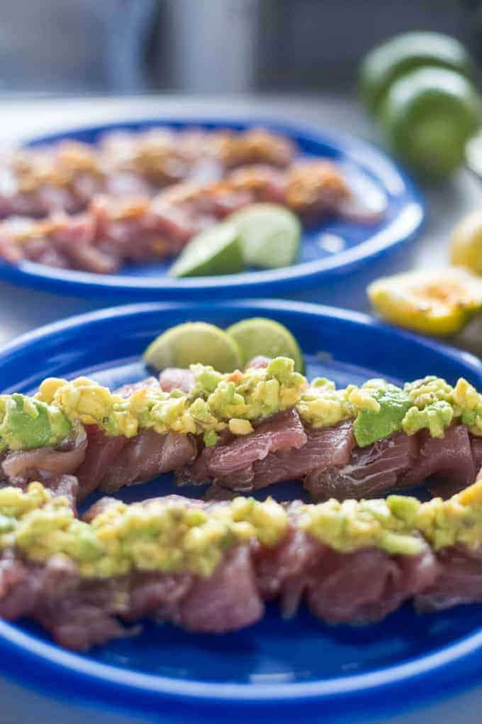 Two blue plates hold rows of raw fish topped with diced avocado and corn. Lime wedges are placed on the plates, with additional limes and lemons in the blurred background.