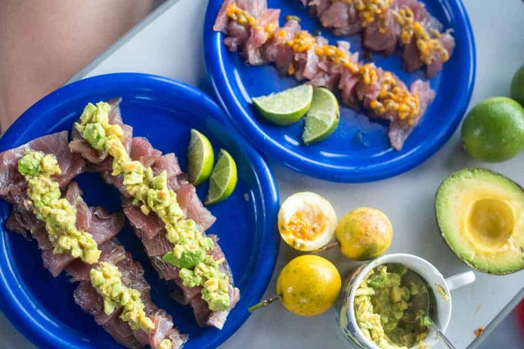Two blue plates hold raw fish topped with mashed avocado and a yellow sauce, with lime wedges on the side. Nearby are halved limes, an avocado, passion fruits, and a mug with leftover mashed avocado.