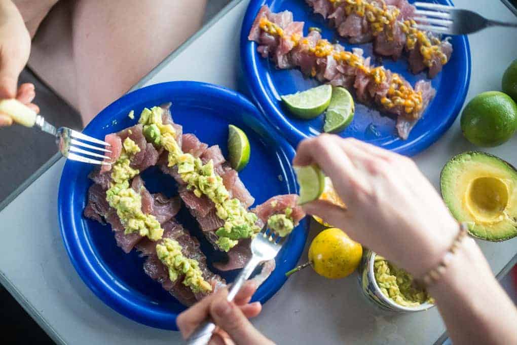 Two blue plates with rows of raw fish topped with guacamole and another topping, served with lime wedges. Hands are seen holding forks and squeezing lime over the fish. Whole avocados and limes are nearby.