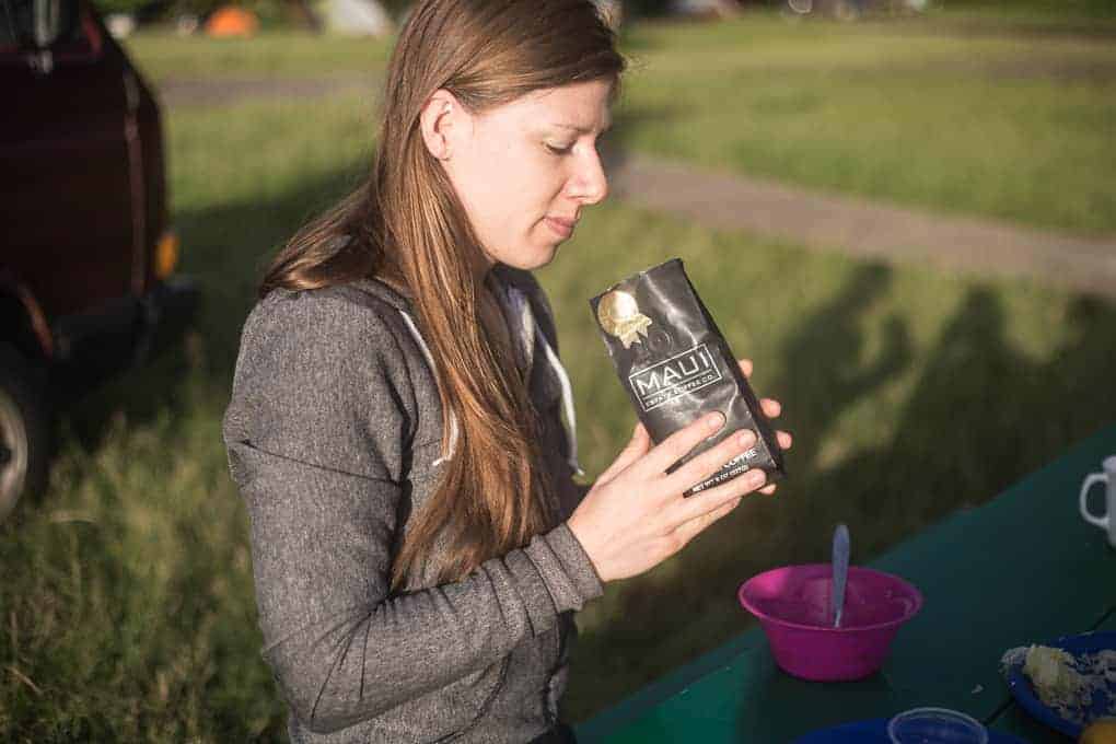 A woman with long brown hair sits at an outdoor table, holding a bag labeled “Maui Coffee Roasters.” A pink bowl, a spoon, and a white mug are on the table. Green grass and a red vehicle are in the background.