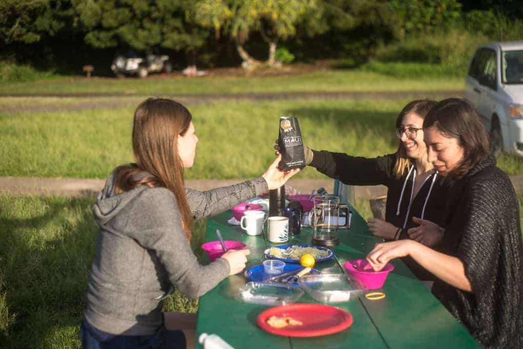 Three women sit at a green picnic table outdoors with food, drinks, and cups. One woman holds up a bag of coffee while the others smile. The background includes grass, trees, and a parked car.