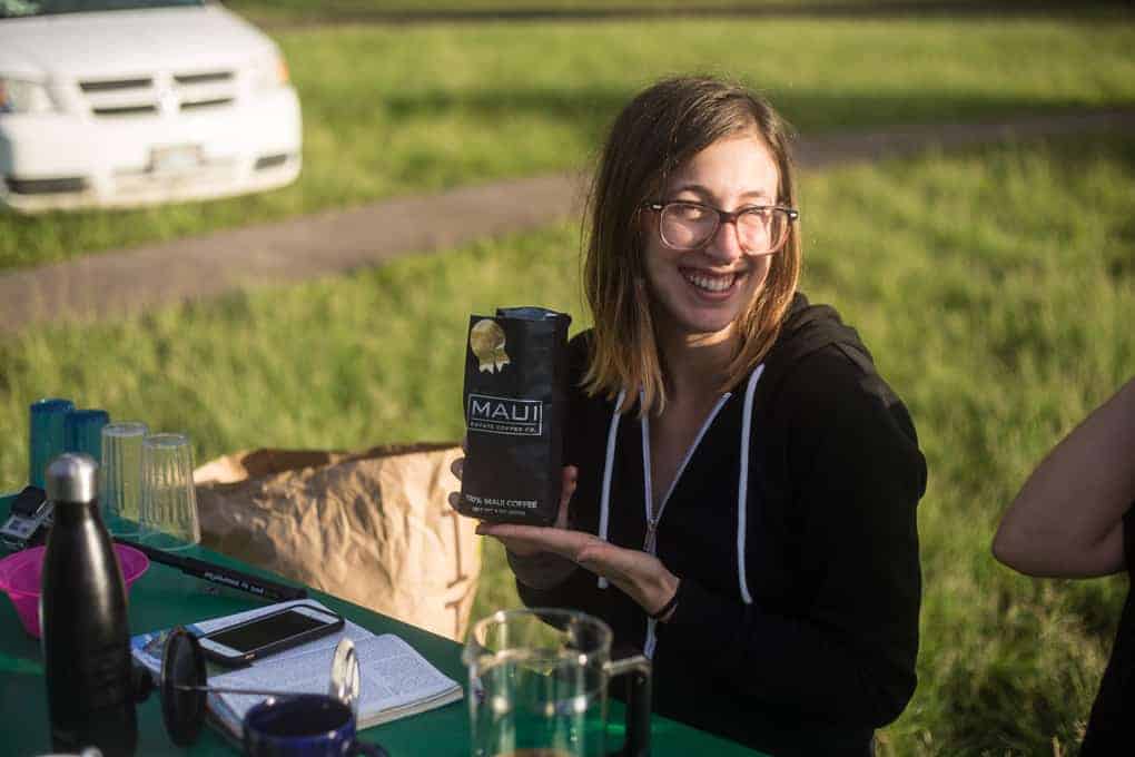 A person with glasses and a black hoodie sits at a green table outdoors, smiling and holding a bag labeled "Maui." Various mugs, bottles, and notebooks are on the table. Grass and a white car are in the background.