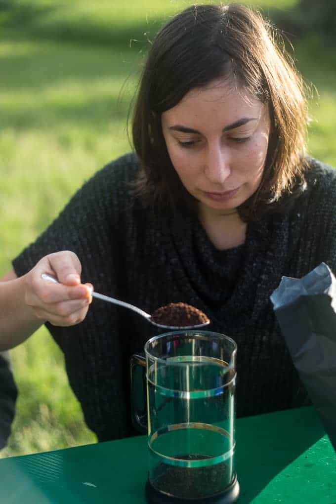 A person with shoulder-length brown hair holds a spoonful of ground coffee over a French press on a green outdoor table, with sunlight and grass in the background.