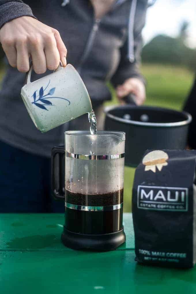 A person pours hot water from a ceramic mug into a French press with coffee grounds on a green table. A bag of Maui coffee sits next to the French press, and a saucepan is held in the background.