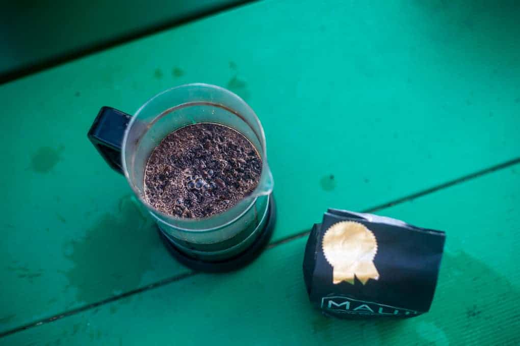 A French press with coffee grounds sits on a green surface next to a black bag of coffee labeled "MAL" with a gold ribbon design.