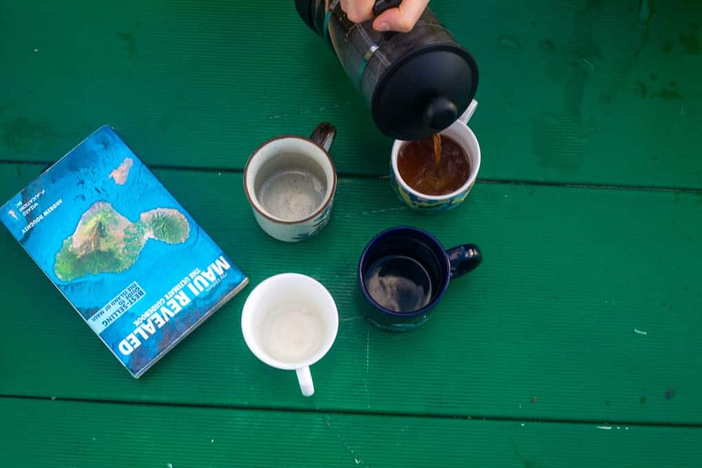 A person pours coffee from a French press into a mug. Three empty mugs and a book titled "Maui Revealed" are arranged on a green surface.