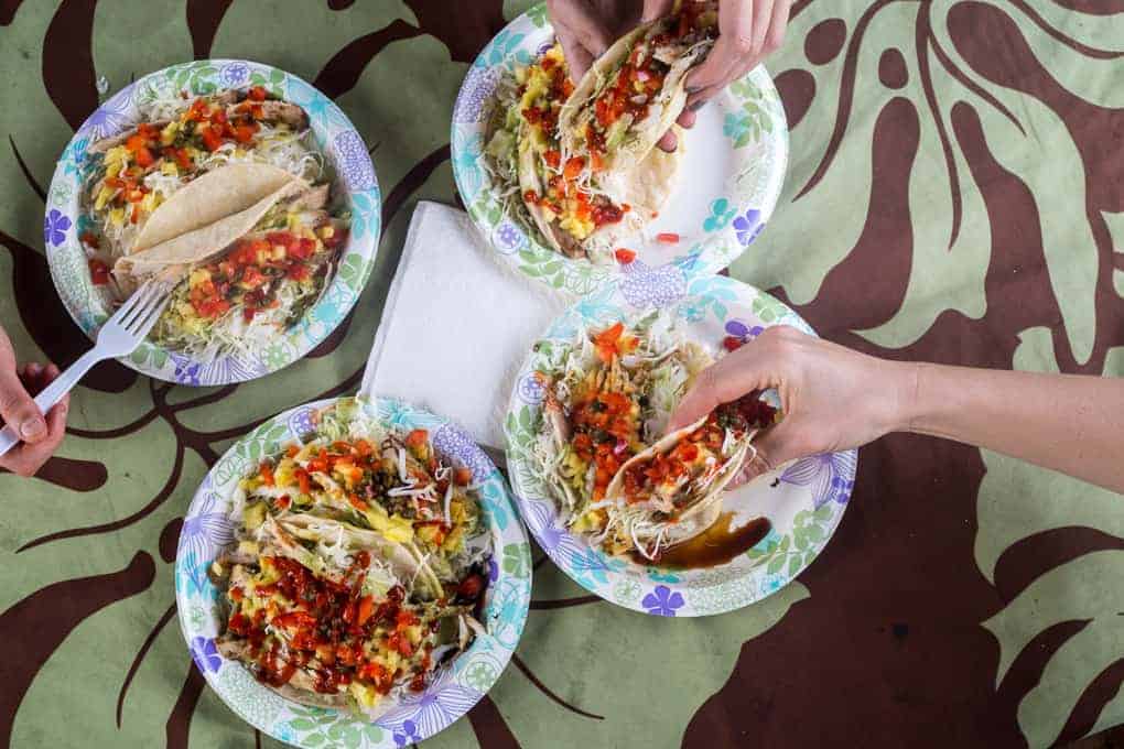 Four paper plates with tacos filled with lettuce, tomatoes, and other toppings are on a table with a floral tablecloth. Two hands are holding tacos, and another hand is using a fork. A napkin is in the center.