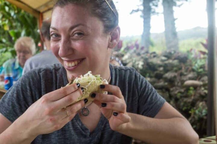 A woman with pulled-back hair and dark nail polish smiles while holding a taco at an outdoor table. There are people and trees in the blurred background.