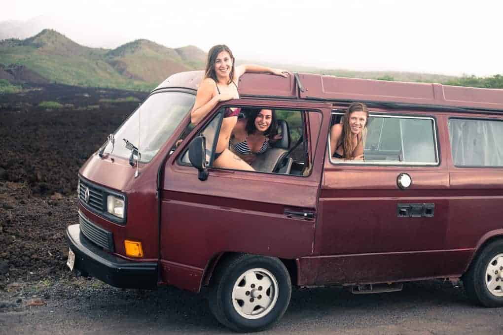 Three women in swimsuits pose and smile from the open windows and door of a maroon van parked on a dirt road, with green hills and vegetation visible in the background.