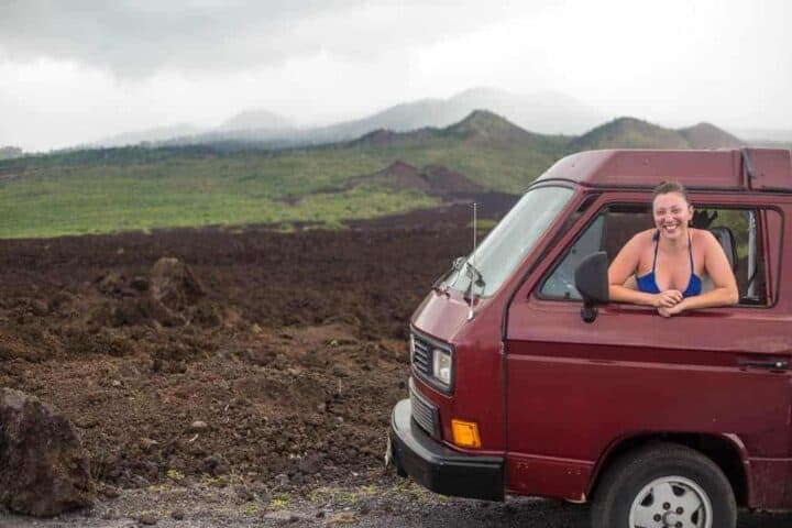 A woman in a blue bikini top leans out of the window of a maroon van, smiling at the camera. The van is parked near rocky terrain with mountains and cloudy skies in the background.