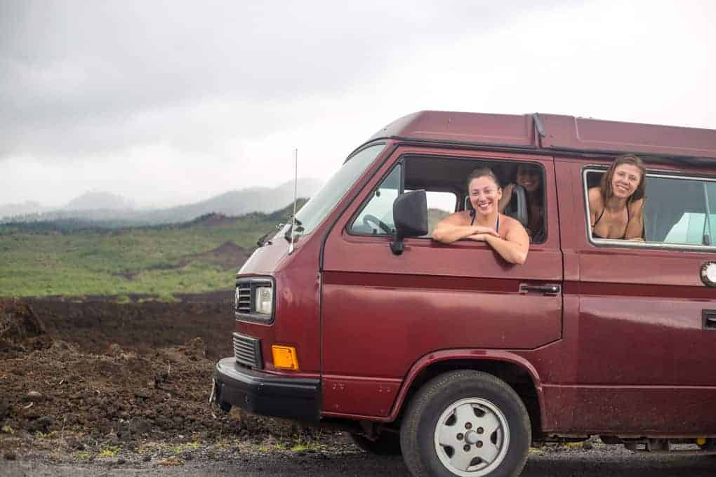 Three people lean out of the windows of a maroon van parked on a rocky roadside with green hills and cloudy sky in the background.
