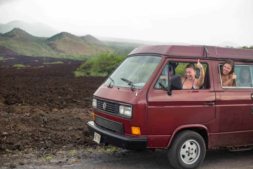 A maroon van is parked on the side of a road with volcanic rock and hills in the background. Three people inside the van smile and give thumbs up out of the open windows.