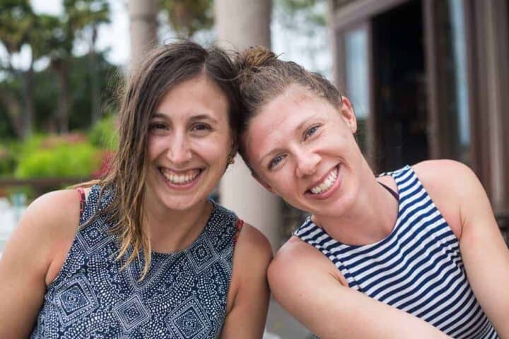 Two women sit closely together outdoors, both smiling at the camera. One wears a patterned sleeveless top, and the other wears a sleeveless striped shirt. Greenery and a building are visible in the background.