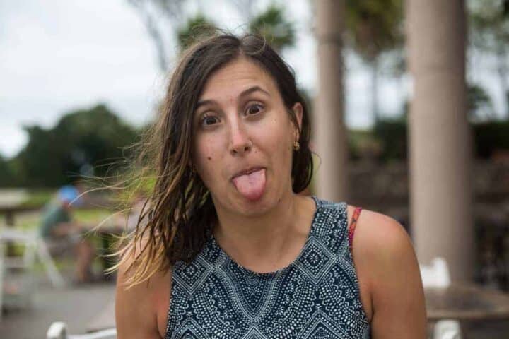 A woman with long brown hair wearing a patterned sleeveless top sticks out her tongue and looks at the camera. There are blurred trees and columns in the background.