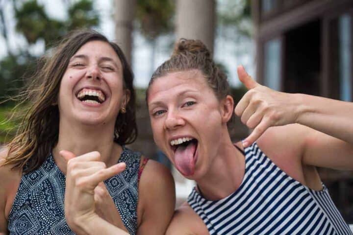 Two women smiling and posing for the camera, one with her tongue out and both making hand signs. They are outdoors, and the background is slightly blurred with trees and a building visible.