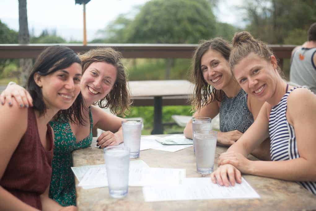 Four women sit at an outdoor table with menus and glasses of water, smiling at the camera. The background includes greenery, a wooden railing, and an out-of-focus person seated behind them.