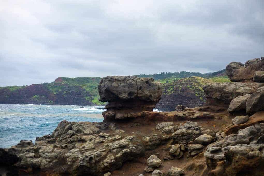 Large rock formation resembling a human head sits near the shoreline, with choppy blue ocean waves, rocky ground, and green-covered cliffs under an overcast sky in the background.