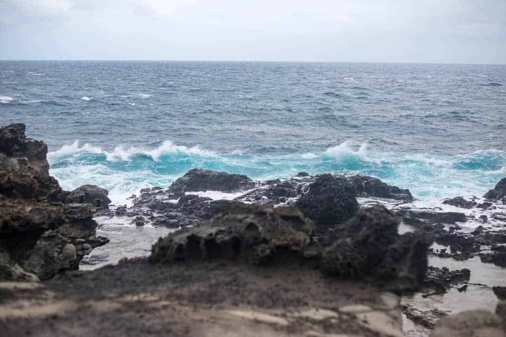 Rocky shoreline in the foreground with waves crashing against dark rocks under a cloudy sky, and the ocean extending to the horizon.
