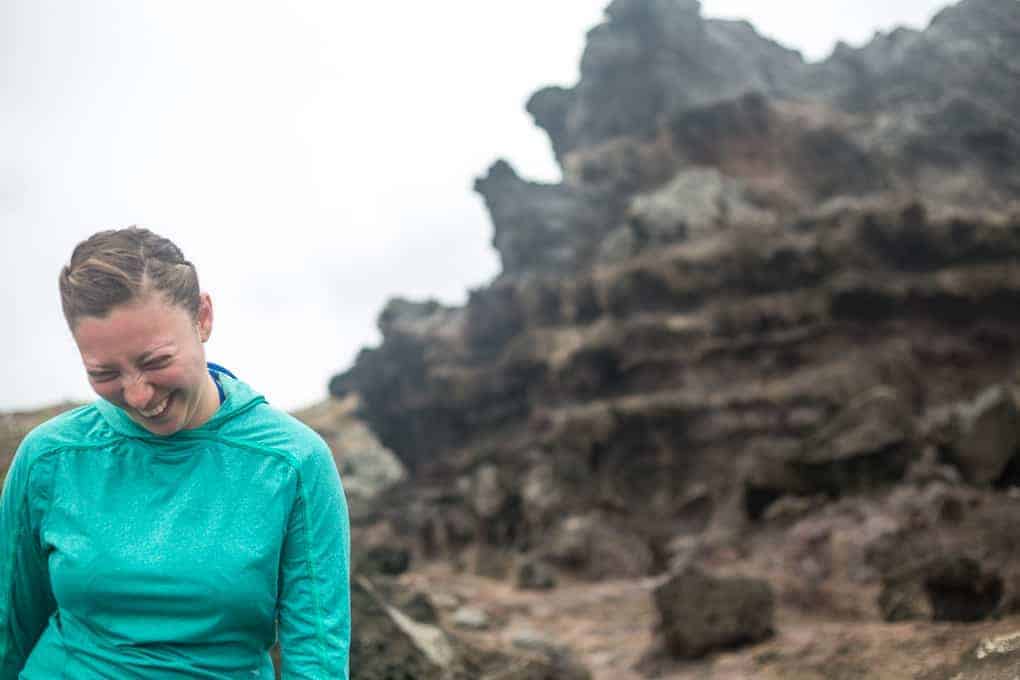 A person in a teal hoodie stands outdoors, looking down and smiling with eyes closed. There are large, rocky formations in the background under an overcast sky.