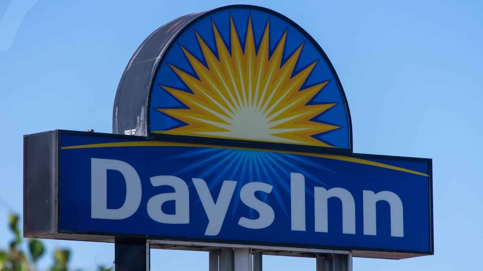 A Days Inn hotel sign with a blue background, yellow sunburst graphic, and white "Days Inn" text stands against a clear blue sky—a familiar sight among US hotel chains disappointing guests.