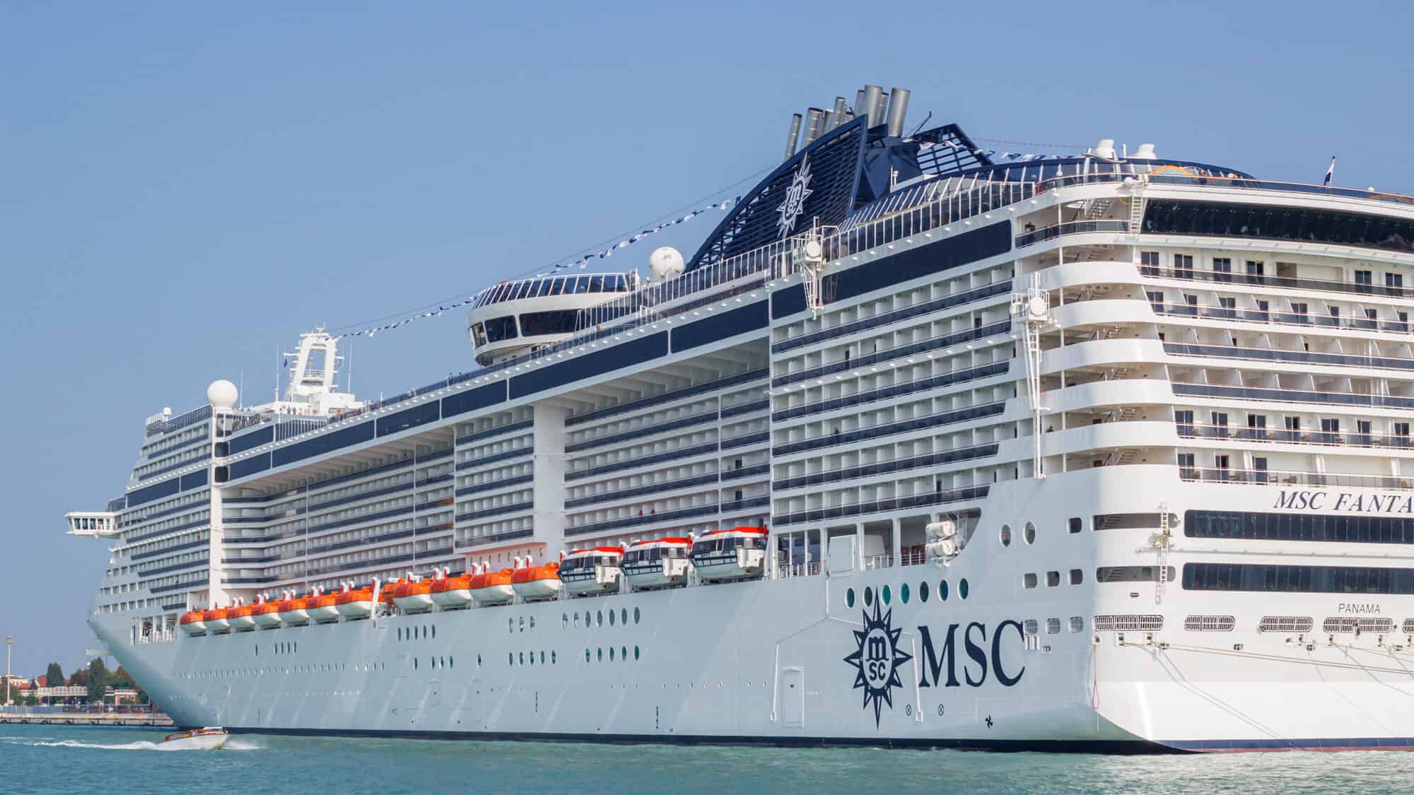 The large white MSC Fantasia cruise ship is docked in calm blue water under a clear sky, with multiple decks, lifeboats, and balconies visible—perfect for a 2025 voyage or Cruise Review.