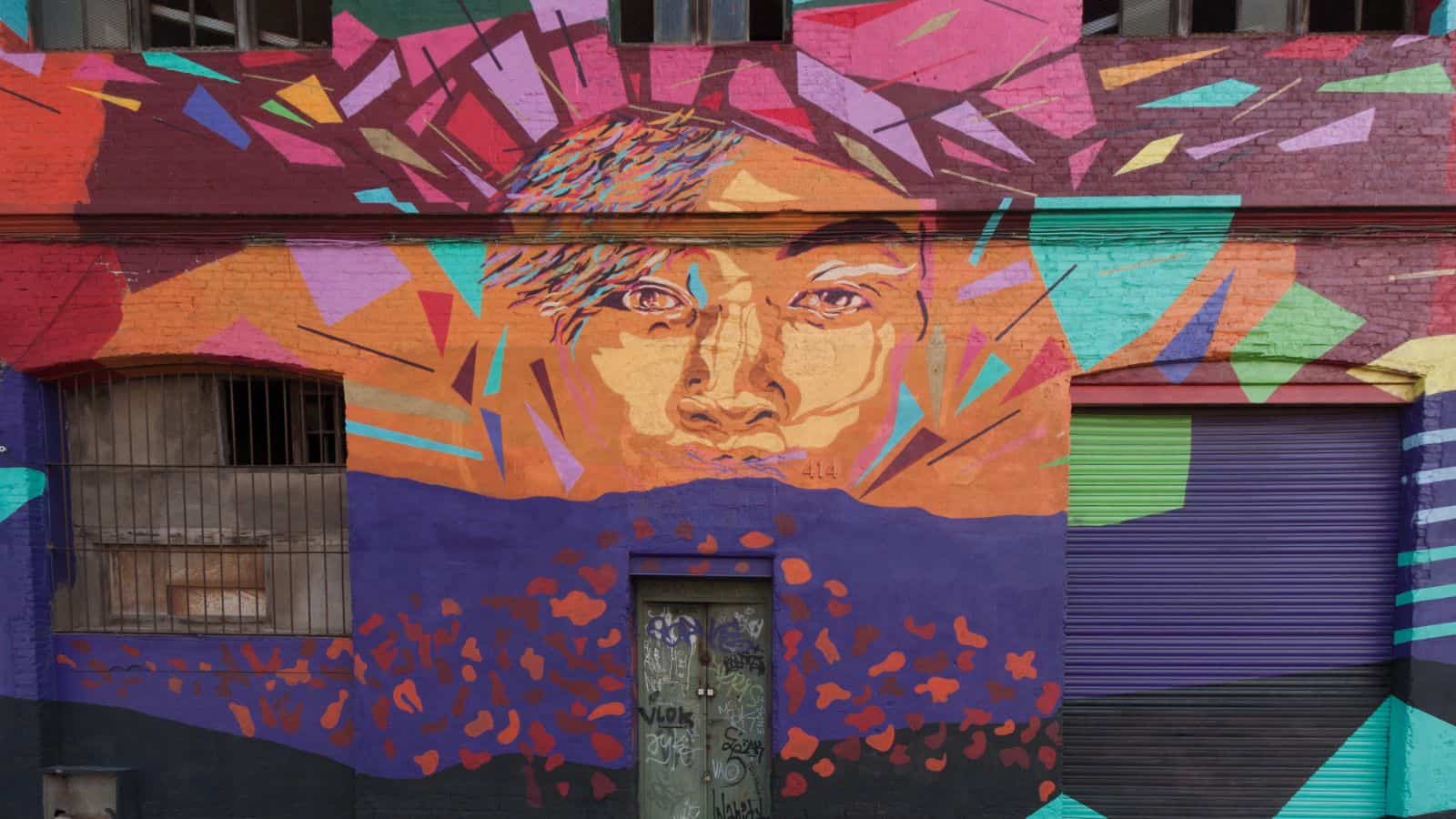 A colorful mural on a brick building features a stylized face surrounded by geometric shapes in shades of orange, purple, red, and teal. The mural covers a door, a window, and a garage-style door.