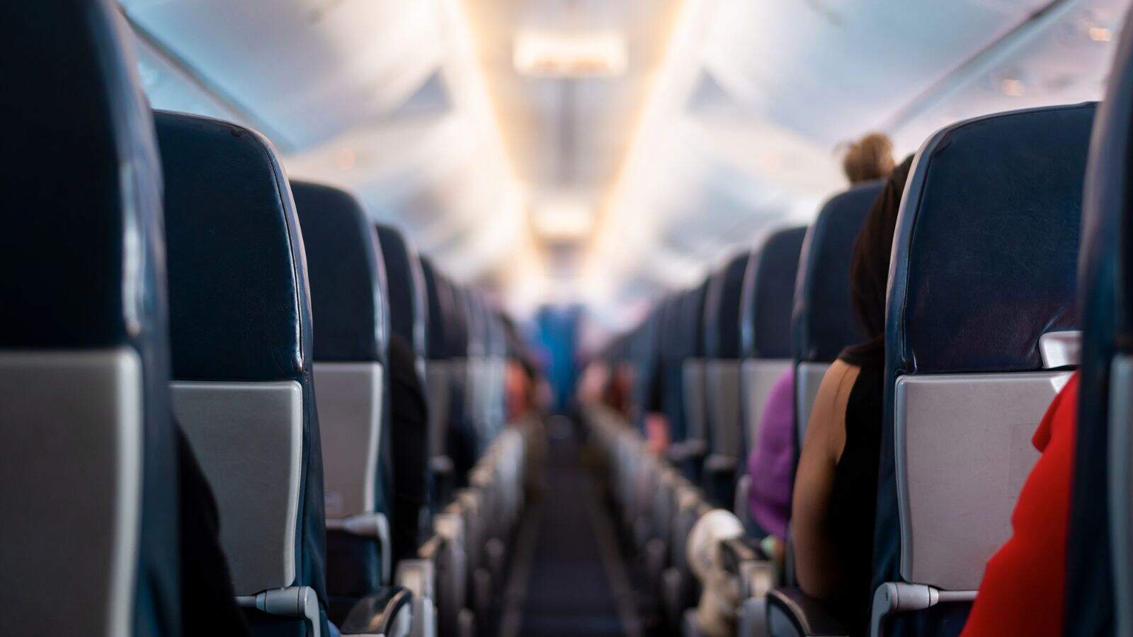 A view down the aisle of an airplane cabin, showing rows of dark seats and passengers seated on either side, with some people visible from behind. The lighting is bright and the focus is on the middle of the aisle.