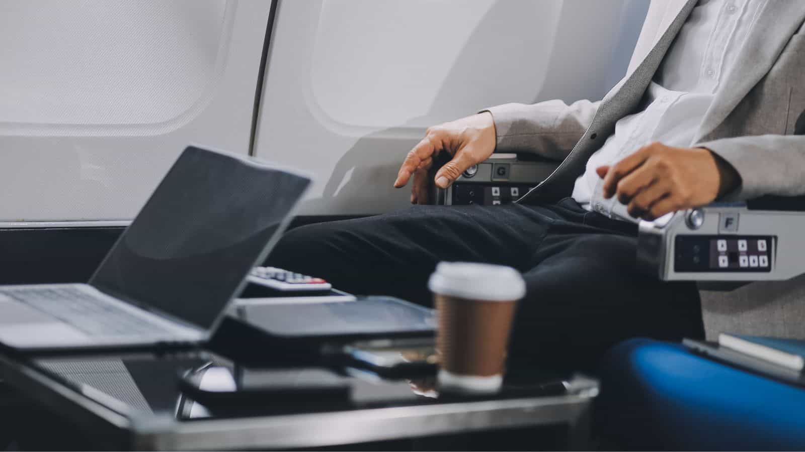A person in business attire sits in an airplane seat next to a tray table holding a laptop, a smartphone, some papers, and a disposable coffee cup. The cabin wall and airplane window are visible in the background.