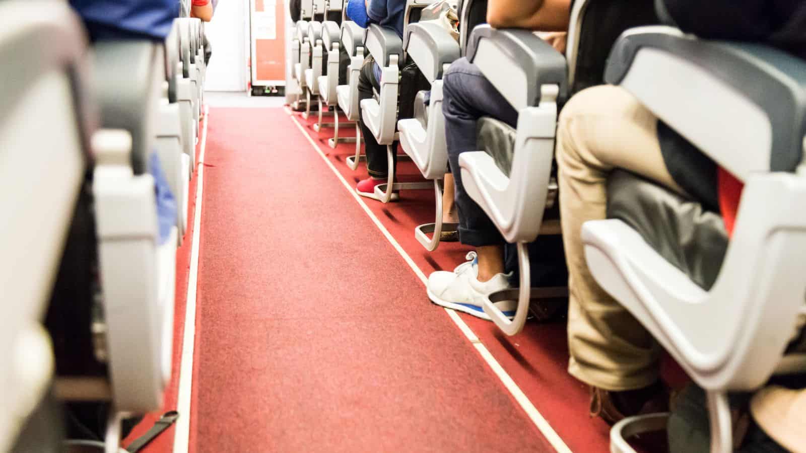 A view down the aisle of an airplane cabin, showing rows of passenger seats on both sides and several people seated. The floor is covered with red carpet, and a pair of white sneakers is visible under one seat.