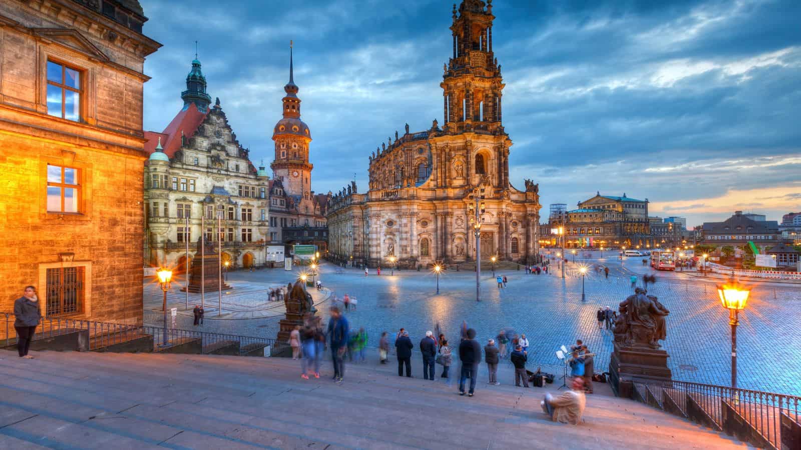 People stand on stone steps overlooking a historic European square with ornate buildings, statues, street lamps, and a large church under a blue evening sky, creating a vibrant urban scene.