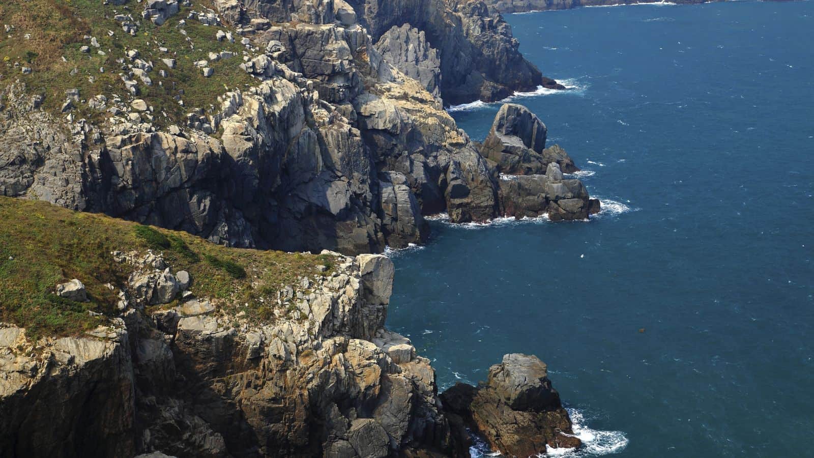 Steep rocky cliffs covered in patches of grass border a deep blue ocean, with waves gently hitting the rocks. The coastline curves in the distance, and the sky is not visible in the image.