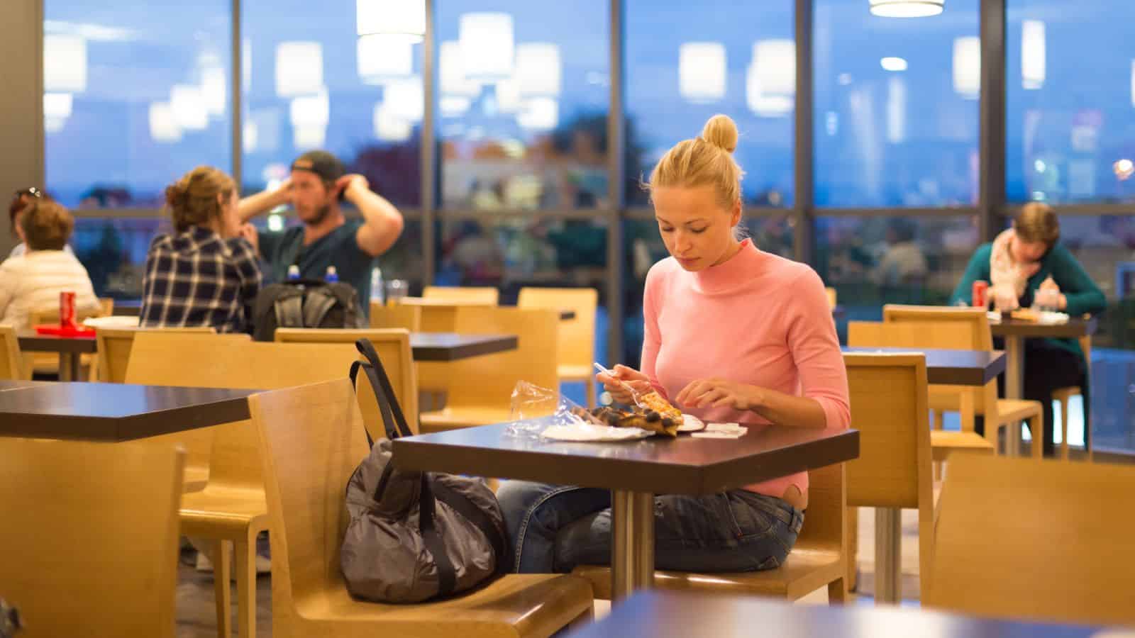 A woman, looking weary from a long flight, sits alone at a table in a brightly lit cafe, eating food from a plastic container. Other people chat in the background as large windows reveal an evening city view.