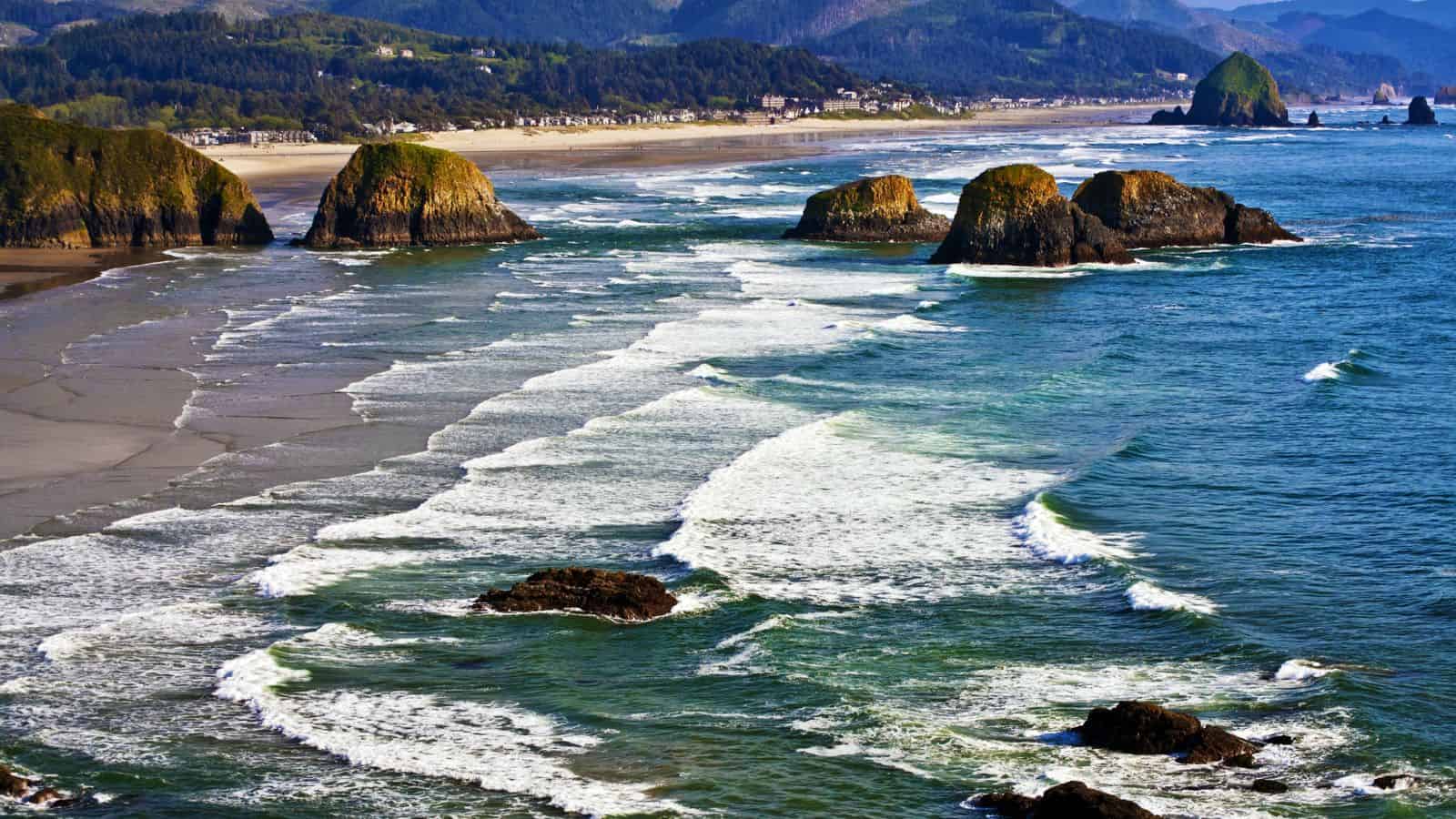 Waves roll in toward a sandy beach with large rock formations in the water; a coastal town and forested mountains appear in the background under a clear sky.