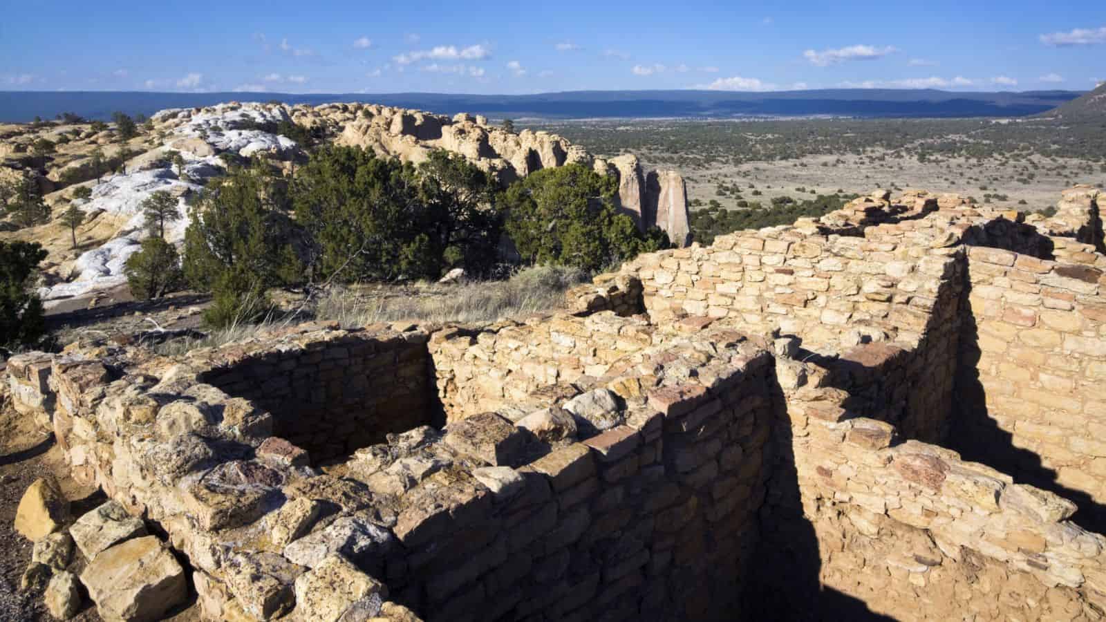 Stone ruins of an ancient structure sit atop a hill with sparse trees and rocky outcrops; a wide, open landscape and distant mountains are visible under a blue sky with scattered clouds.