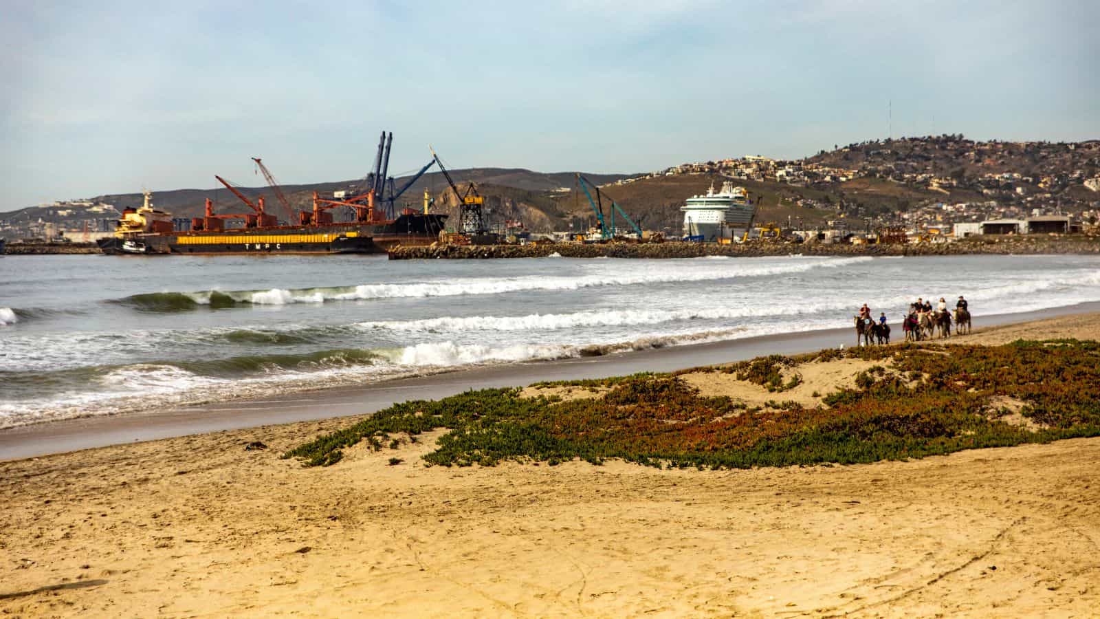 A sandy beach with a few people riding horses near the shore. In the background, a cargo ship and cranes are visible at a port, with hills and buildings further in the distance.