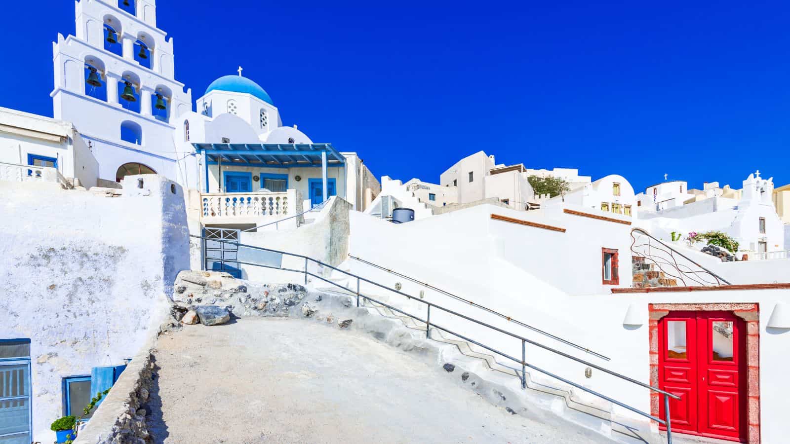 A hillside scene in Santorini, Greece, featuring whitewashed buildings, a church with a blue dome and bell tower, a sloped walkway with railings, and a red double door under a clear blue sky.