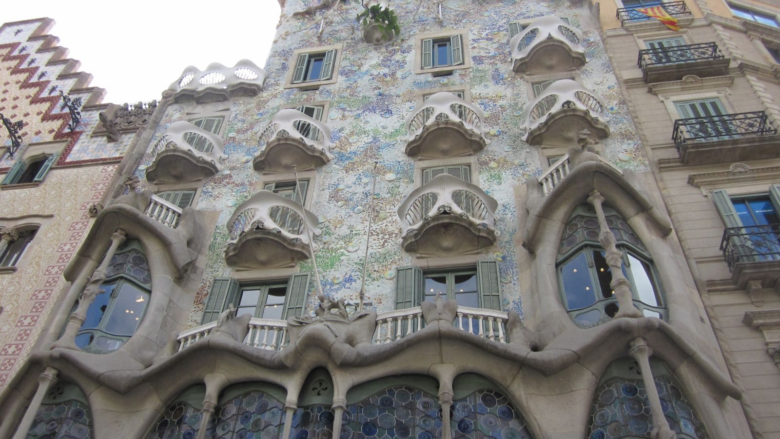 The image shows the ornate facade of Casa Batlló in Barcelona, featuring curved windows, balconies resembling masks, and colorful mosaic tiles, with some neighboring buildings partially visible on both sides.