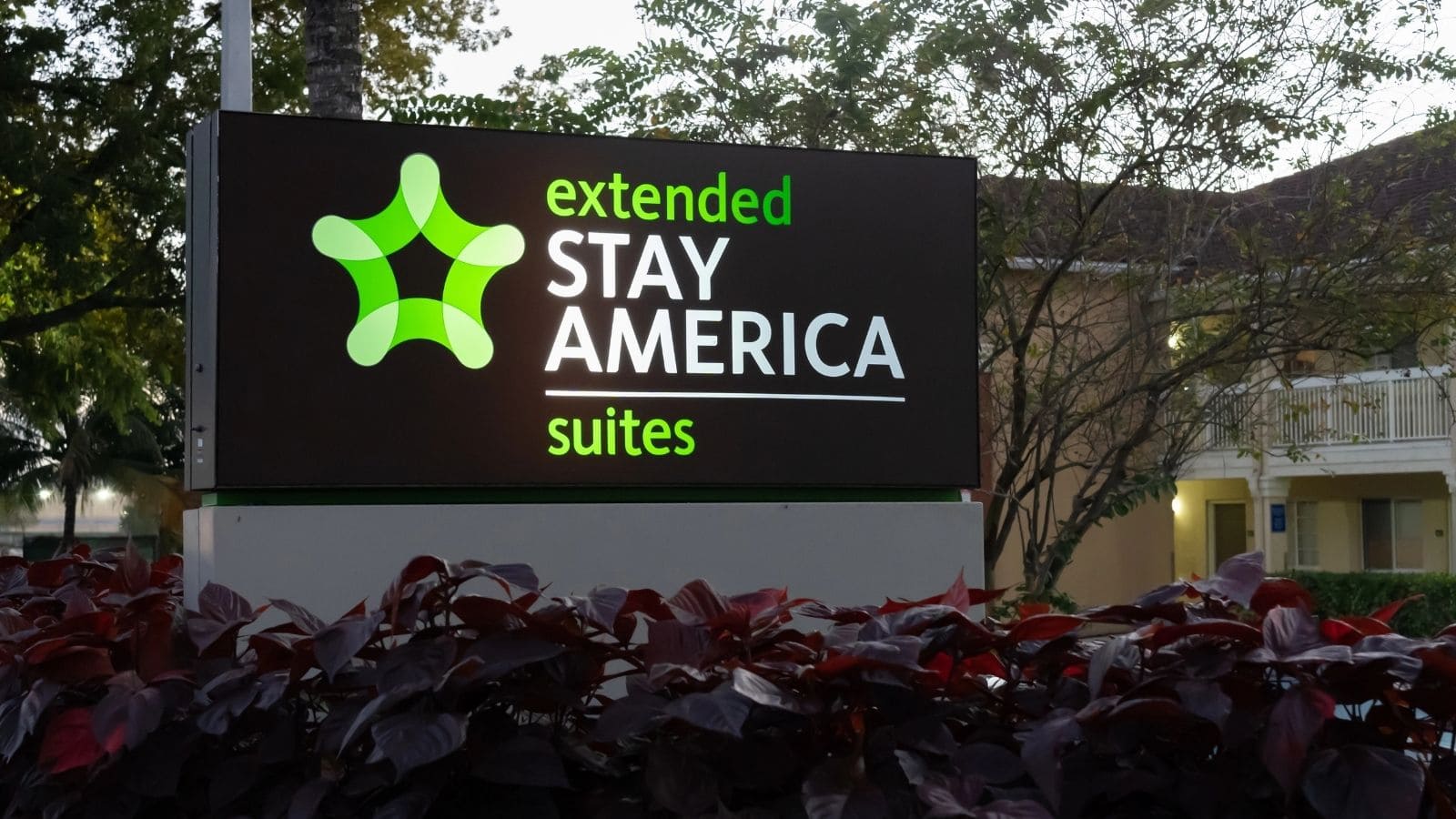 A sign reads “extended STAY AMERICA suites” in green and white letters, with a green logo above the text. Bushes and trees are in the foreground and a building is visible in the background—one of several US hotel chains disappointing guests.