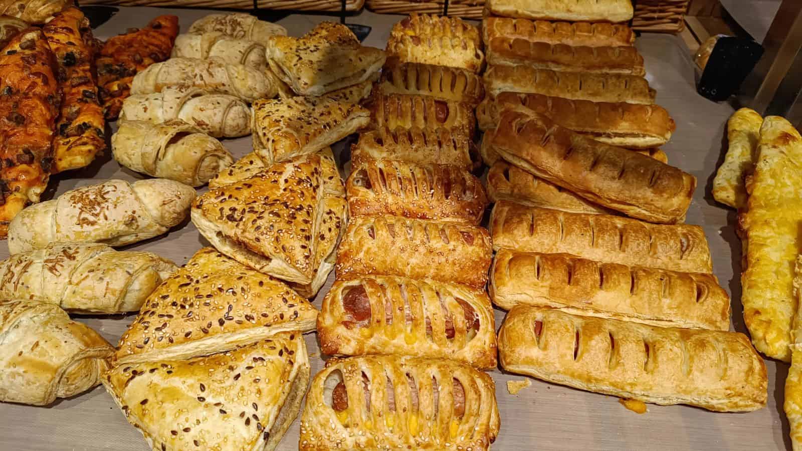 An assortment of baked goods, perfect for all types of travelers, including puff pastry triangles with seeds, sausage rolls, and rectangular pastries arranged on a wooden surface. Wicker baskets are visible in the background.