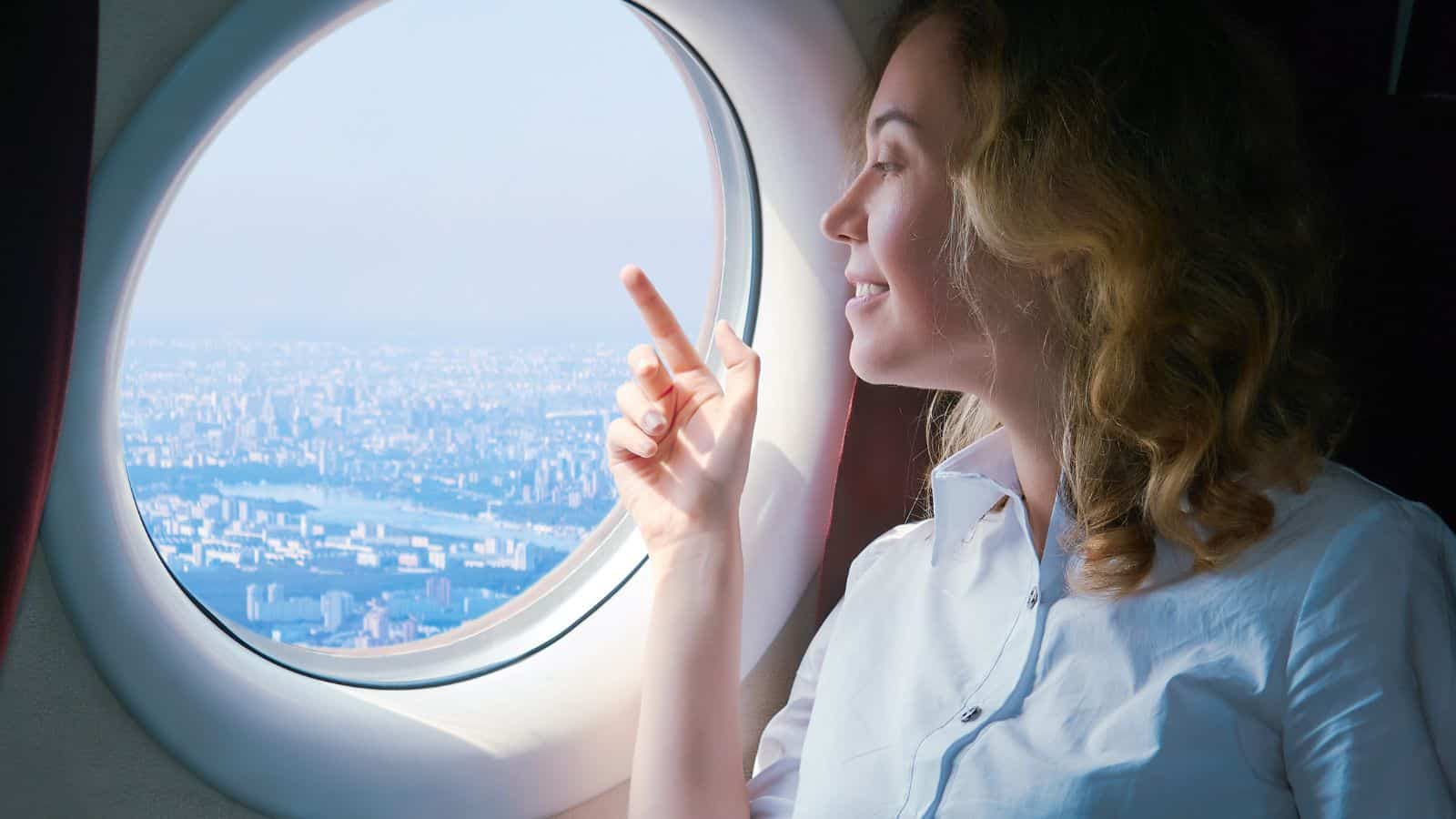 A woman with wavy hair and a white shirt sits by an airplane window, smiling and pointing outside at the cityscape below&mdash;proof that it&rsquo;s possible to conquer fear of flying and enjoy the journey.