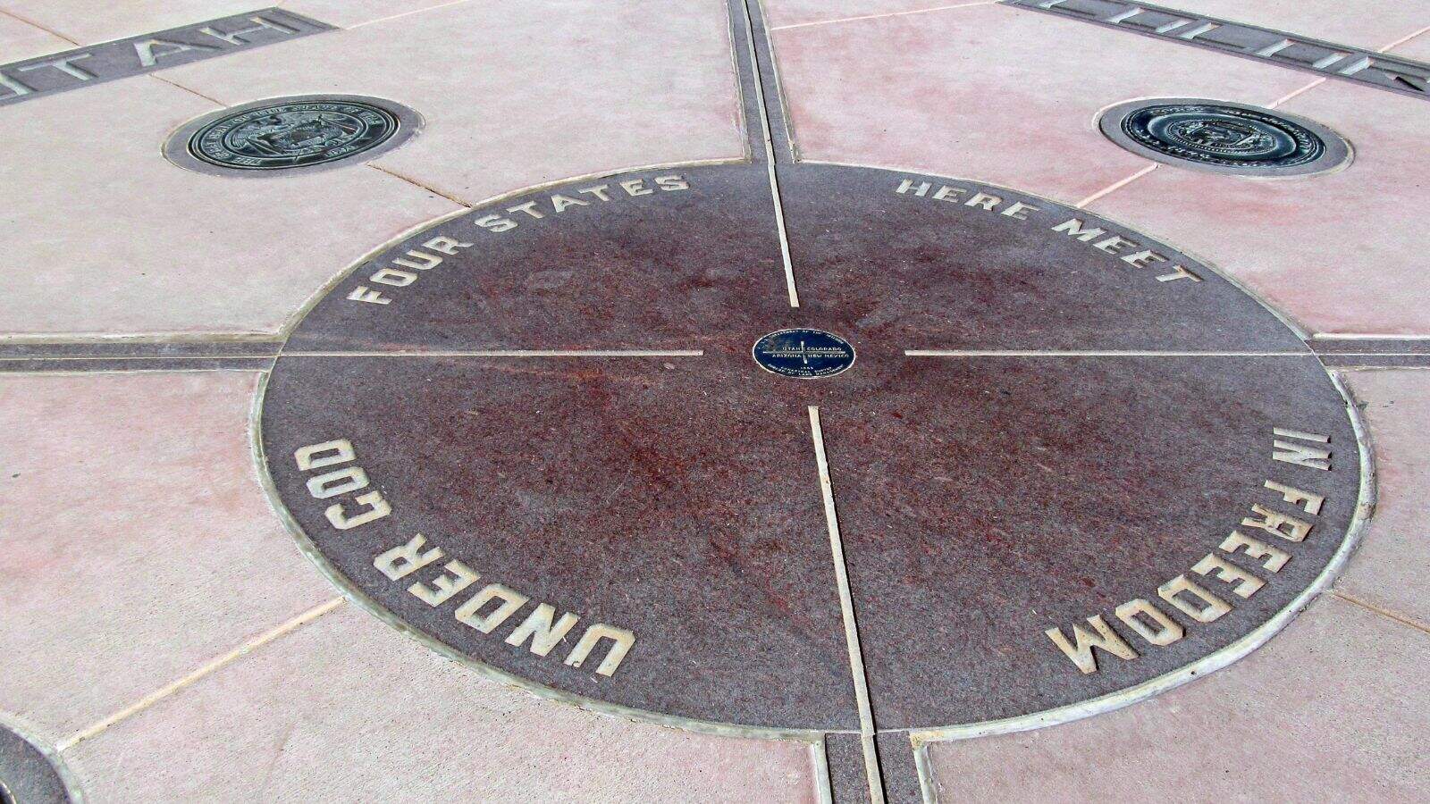 A circular monument marks the point where four U.S. states meet—Utah, Colorado, Arizona, and New Mexico—with state seals and engraved text stating “Four States Here Meet” and “Under God In Freedom.”.