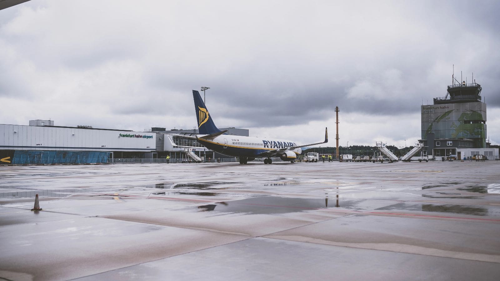 A Ryanair airplane is parked on a wet airport tarmac near a terminal building and a control tower under a cloudy sky. The terminal has signage in German, and the ground is reflective from recent rain.